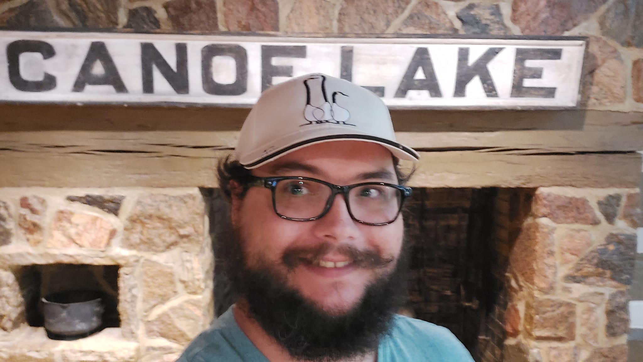 A man with short hair and bushy facial hair stands in front of a stone fireplace with a sign reading "Canoe Lake" on the mantle